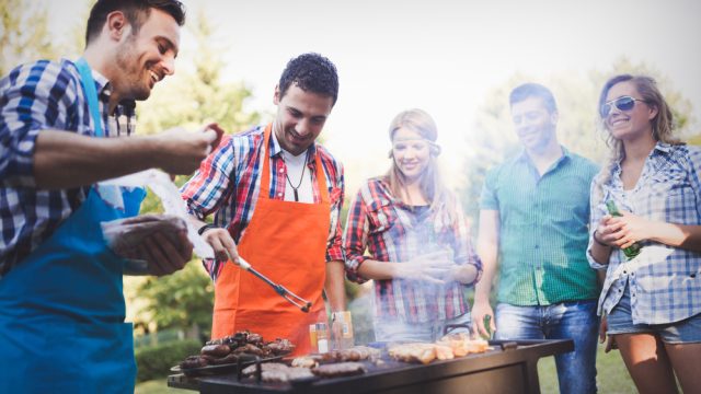 A group of friends standing around a barbecue while meat is grilled