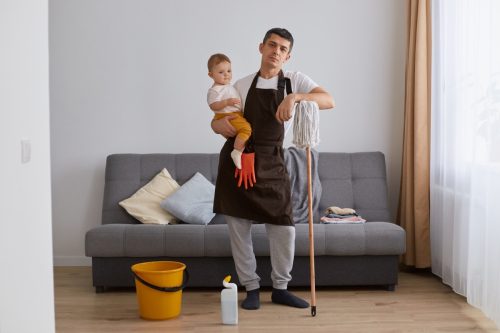 young adult man wearing casual attire and brown apron cleaning house with baby in hands, looking at camera with sad upset facial expression.
