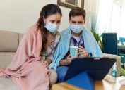 Scared couple in masks sitting on sofa and consulting online with a doctor over digital tablet