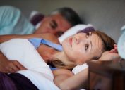 woman looking concerned in bed next to her alarm clock while her husband sleeps next to her