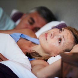 woman looking concerned in bed next to her alarm clock while her husband sleeps next to her