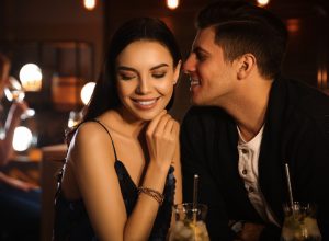 woman smiling seductively while man whispers in her ear at a bar