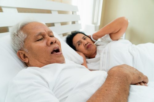 older man snoring in bed while woman covers ears