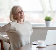 A senior woman sitting at a table holding her back in pain