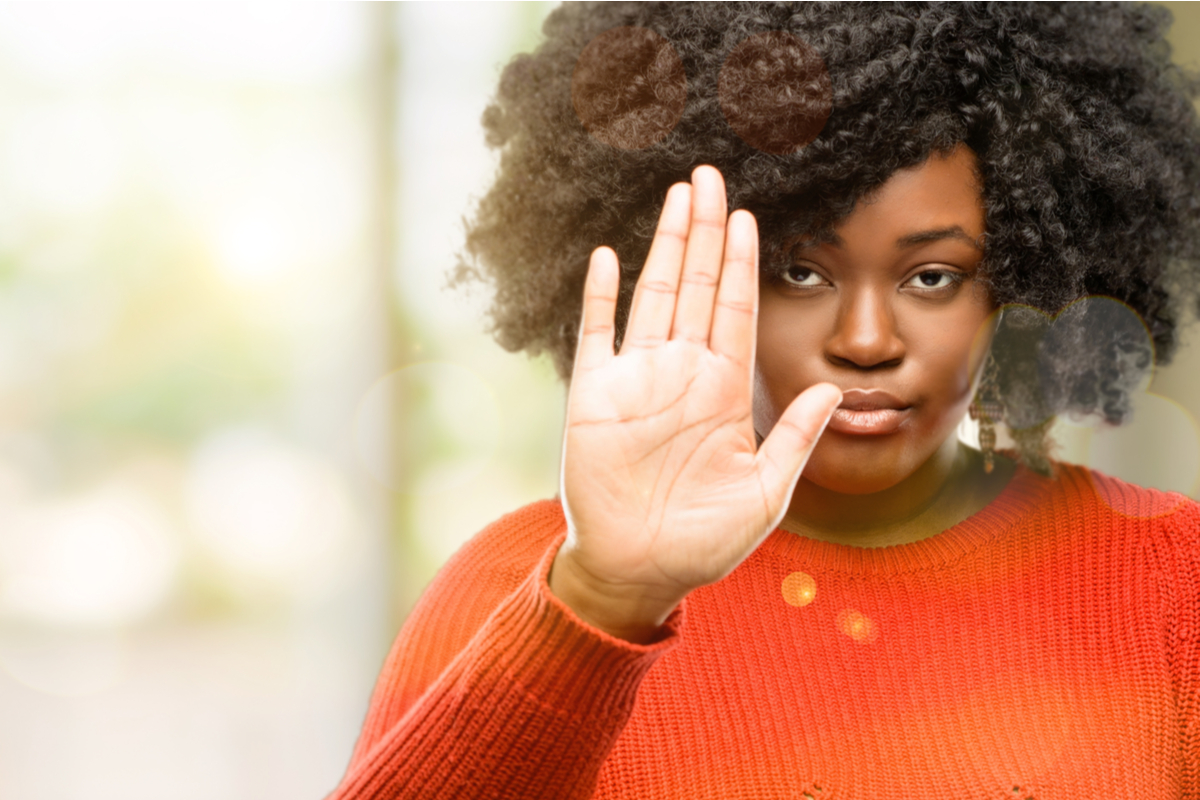 young Black woman wearing orange sweater holding up her hand to say "no"