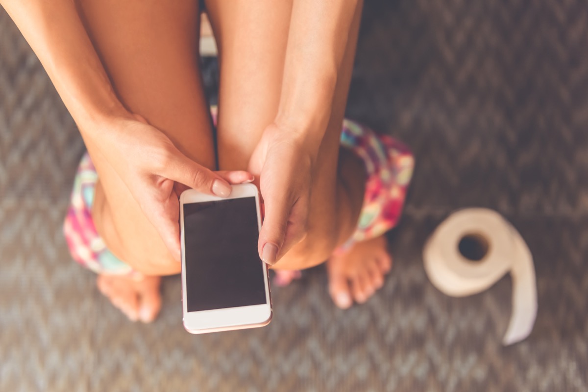 Cropped image of young woman using a smartphone while sitting on toilet