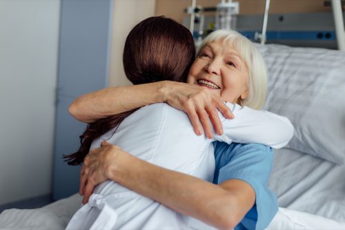 happy senior woman lying in bed and hugging female doctor in hospital