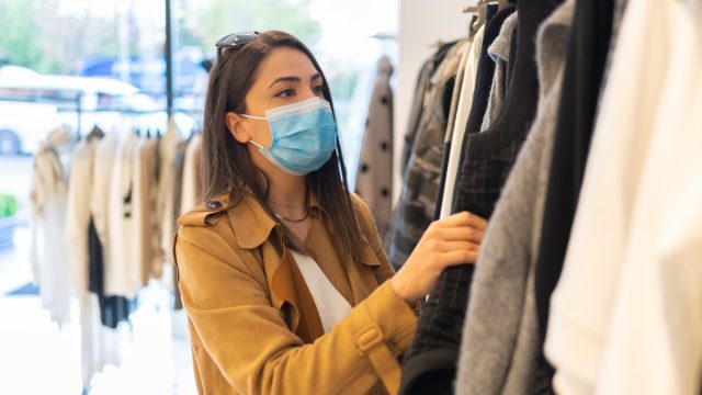 A beautiful young stylish woman with protective face mask is choosing trendy dress in the clothing store during pandemic