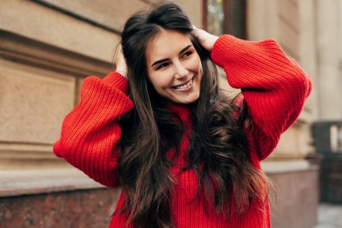 Schöner junger Brunette, der breit mit ihren Händen über ihr langes Haar lächelt.  Außenfoto eines schönen Modells in einer trendigen roten Strickjacke beim Spaziergang durch die Straßen der Stadt.