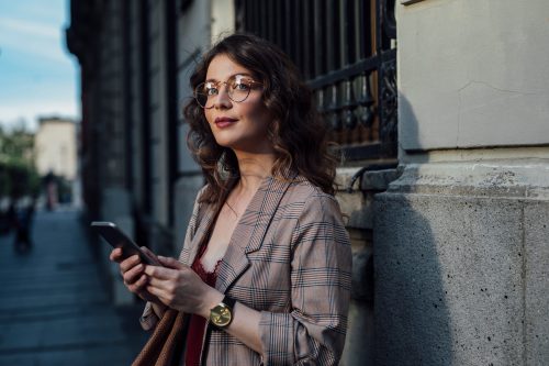 Μια νέα, έξυπνη, κομψή γυναίκα στέκεται στο δρόμο, κρατώντας το τηλέφωνό της.
