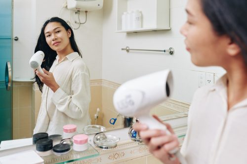Smiling pretty young Vietnamese woman blow drying hair in front of mirror in bathroom