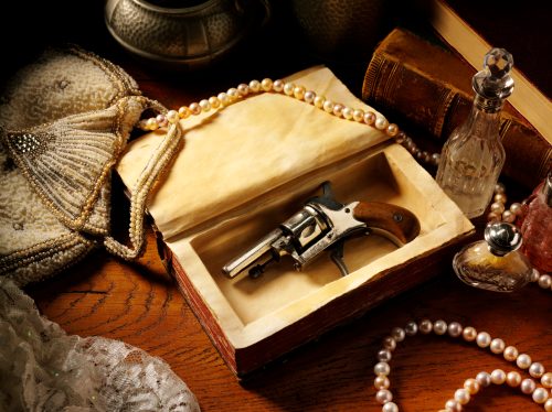 Poză cu tematică vintage cu un pistol edwardian, o sticlă care adulmecă și perle pe o masă antică din lemn.