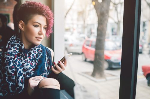 Fată atrăgătoare cu păr scurt, așezată lângă o fereastră într-o cafenea, ținând un smartphone și uitându-se afară
