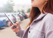 woman holding mobile phone in hand standing near cars parking lot. Searching information and news. Social media concept.