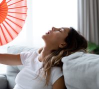 Close up overheated woman waving paper fan, breathing air, leaning back on couch alone, exhausted sick young female feeling unwell, suffering from heating, fever or hot summer weather at home