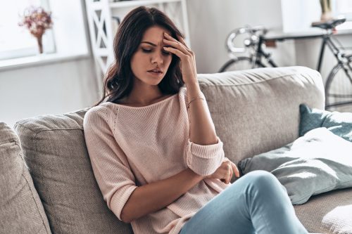 A young woman sitting on the couch holding her head while sick with cold, flu, or COVID symptoms