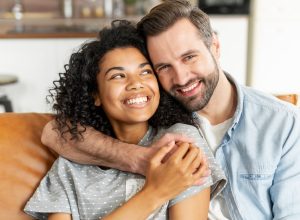 A couple sits on the couch embracing and smiling.