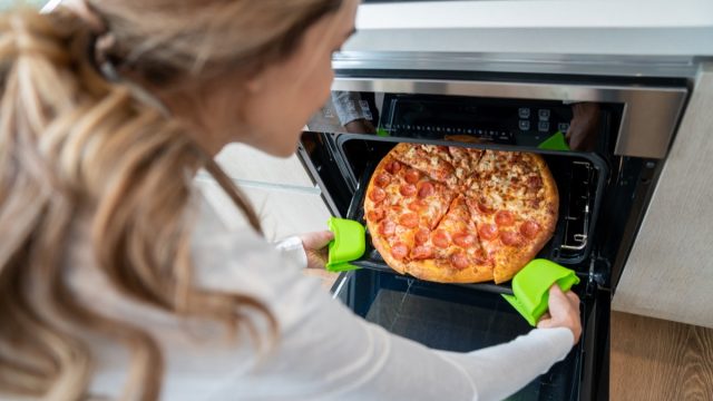 High angle view of unrecognizable woman taking out a fresh baked pizza from oven at home - Lifestyles