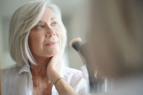 Beautiful senior woman with shiny gray hair in front of mirror with makeup brushes