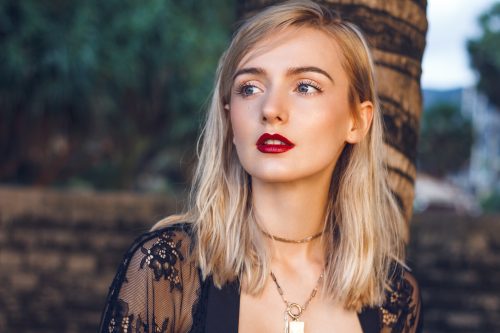 Prim-plan al unei tinere blonde superbe, cu păr drept, ochi verzi misterioși și buze roșii sexy.  Poartă o ținută modernă din dantelă neagră și coliere aurii lângă palmă.