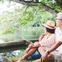 An elderly couple hugging each other with love and happiness in a park with a large pond.