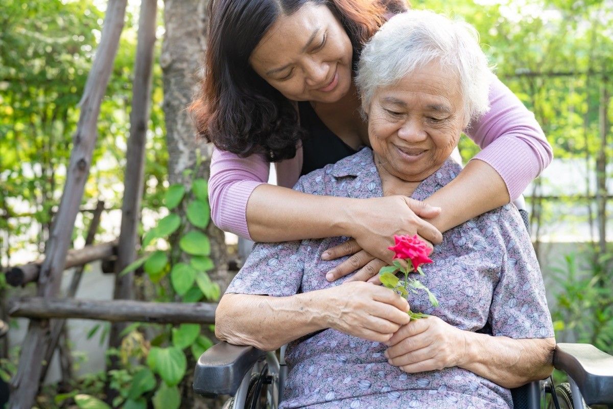 Caregiver daughter hug and help Asian senior or elderly old lady woman holding red rose on wheelchair in park.