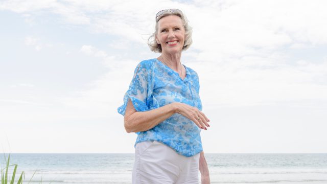 An older woman walking on the beach wearing white shorts and a blue blouse.