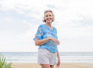 An older woman walking on the beach wearing white shorts and a blue blouse.