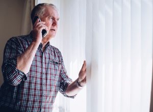 Senior man talking on the phone whilst looking out of his window. He has a worried look on his face.