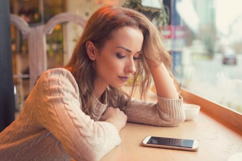 Young woman sitting at table in cafe looking at her phone unhappily.