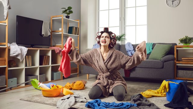 A scattered young woman wearing a face mask and curlers, trying to put away laundry.