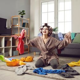 A scattered young woman wearing a face mask and curlers, trying to put away laundry.