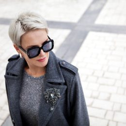 Portrait of fashionable mature woman with short blonde hair wearing stylish gray wool coat with lapels and shoulder straps, accessories brooch and sunglasses