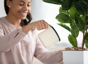 Smiling woman holding a pot watering green plant at home