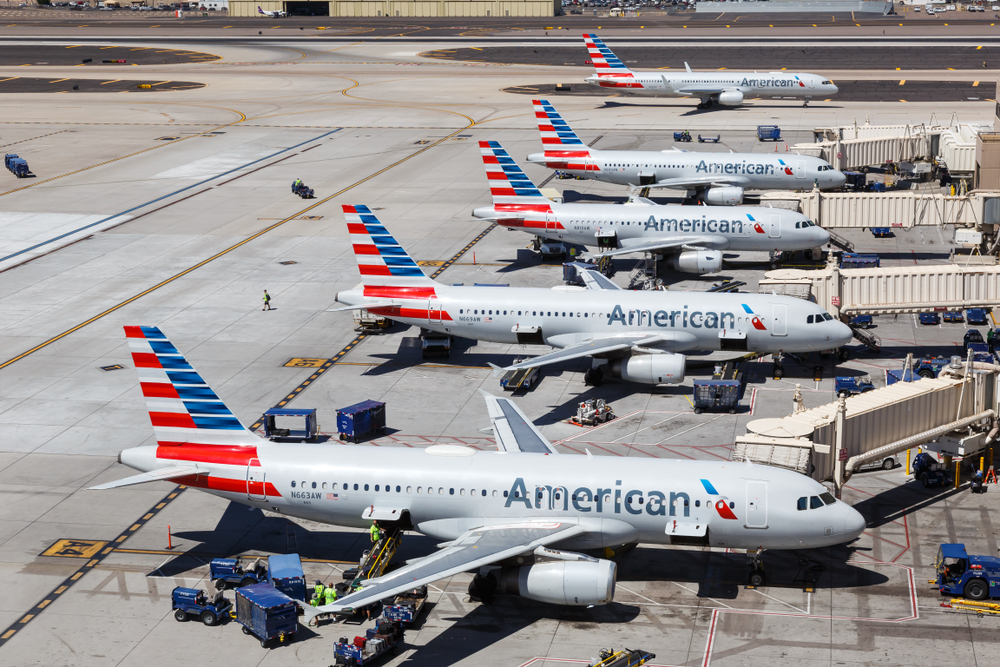 Five American Airlines planes sitting at their gates and one plane taxiing on the runway at an airport