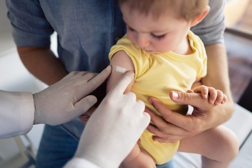 Un medic de nerecunoscut pune un plasture pe umărul unui copil după o vaccinare reușită.  Băiețel care stă în poala tatălui și se uită la plasture.