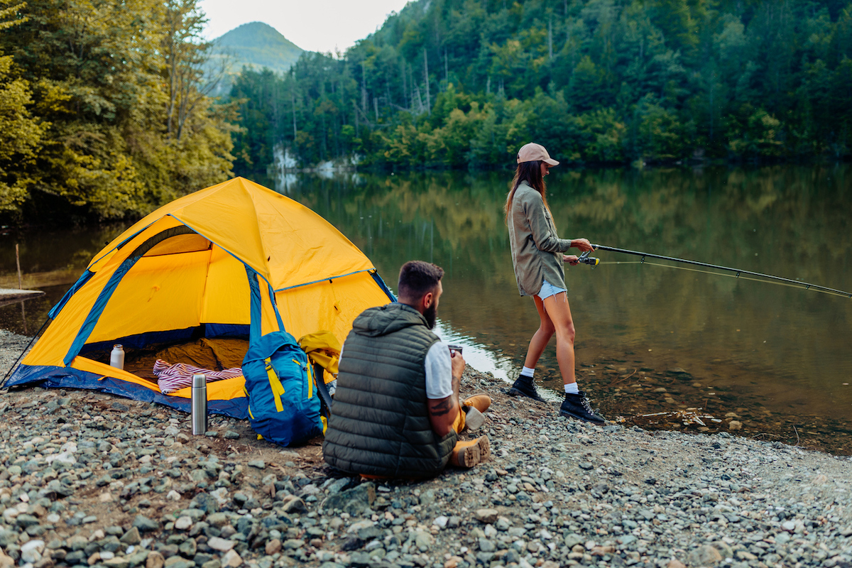 Young woman out fishing by a river while her boyfriend sits next to their yellow camping tent