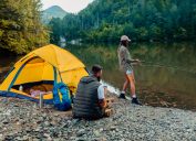 Young woman out fishing by a river while her boyfriend sits next to their yellow camping tent