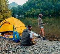 Young woman out fishing by a river while her boyfriend sits next to their yellow camping tent