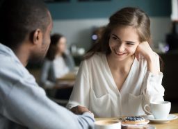 Happy interracial couple flirting talking sitting at cafe table, african man holding hand of smiling caucasian woman having fun drinking coffee together at meeting