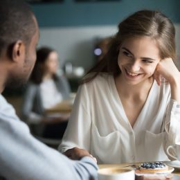 Happy interracial couple flirting talking sitting at cafe table, african man holding hand of smiling caucasian woman having fun drinking coffee together at meeting