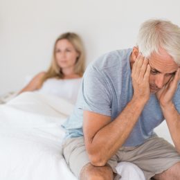 Close up of a worried senior man sitting up in bed with his wife in the background.