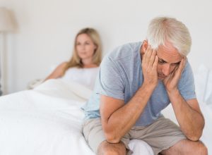 Close up of a worried senior man sitting up in bed with his wife in the background.