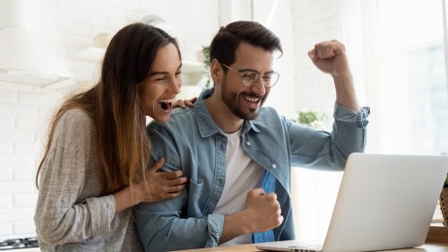A young couple standing over their laptop with a happy expression on their faces and cheering