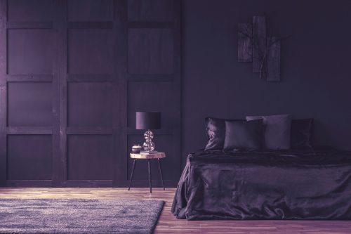O lampă pe o masă de lemn într-un dormitor violet spațios cu lenjerie de pat asortată violet închis