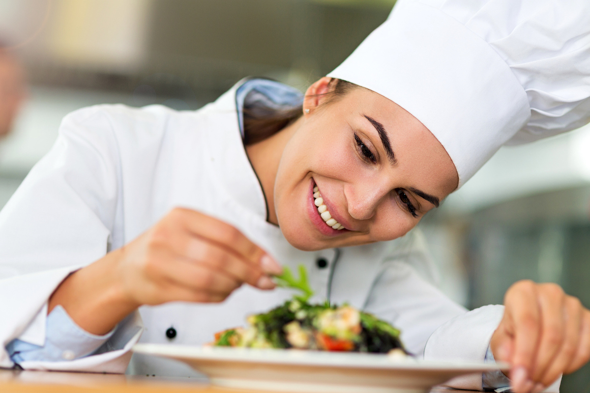 Close up of a female chef in a chef's hat and coat plating a salad