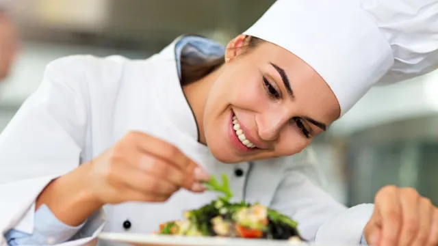 Close up of a female chef in a chef's hat and coat plating a salad