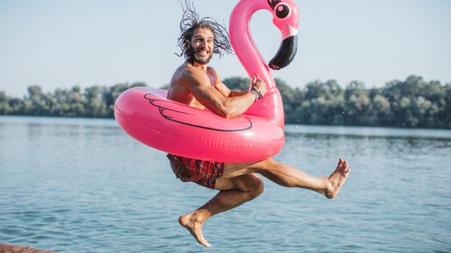 Young man jumping in river from deck. He is jumping with pink flamingo swimming float.