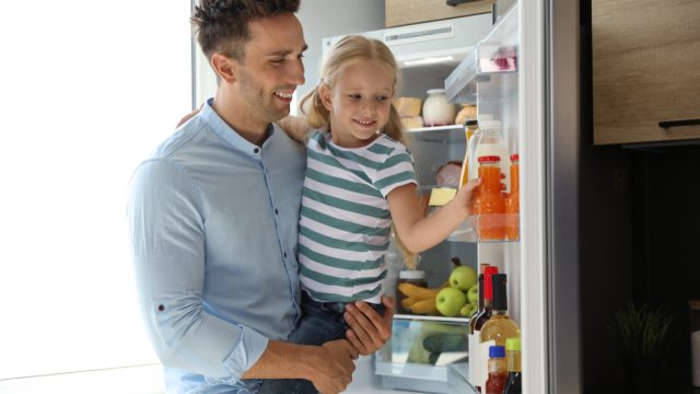 A young girl grabbing a juice out of the fridge while being held up by her father