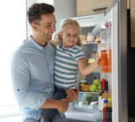 A young girl grabbing a juice out of the fridge while being held up by her father